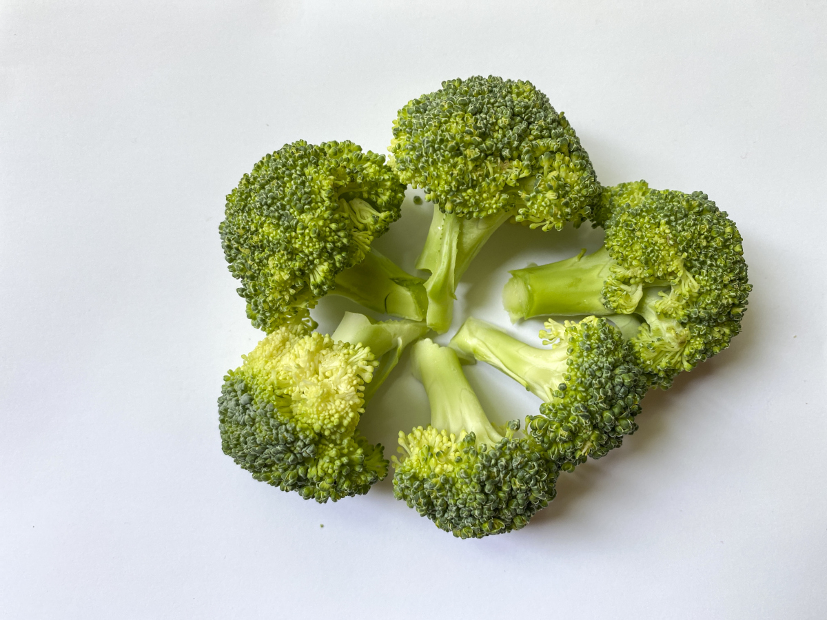 家庭厨房场景：砧板上摆放三种西兰花料理成品，左侧为蒜蓉清炒西兰花（翠绿油亮、花球完整）；中部为橄榄油低温烘烤西兰花（边缘微焦、表面泛琥珀光泽）；右侧为西兰花茎部刨丝拌芝麻酱油汁（白色脆茎丝与黑芝麻形成对比），三道菜置于天然木纹餐盘中，背景为简洁现代厨房台面