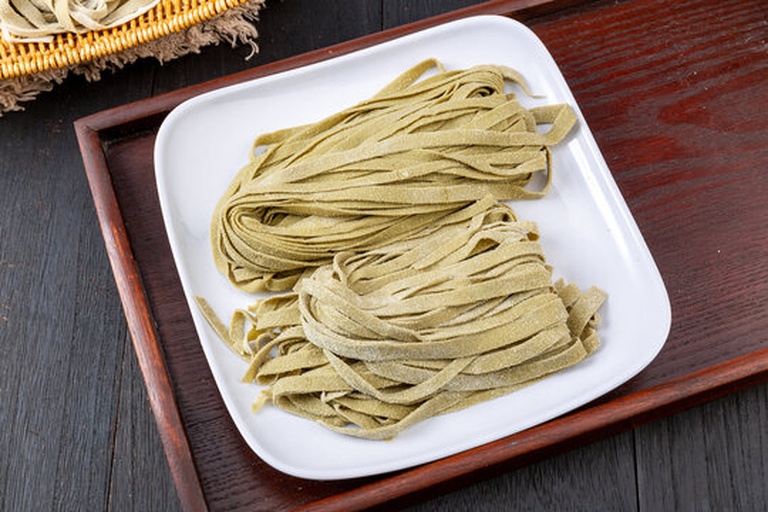 美食摄影布光:粗陶浅碗中盛放荞麦冷面,表面覆盖浓稠翠绿青酱,酱体可见细碎羽衣甘蓝叶脉与金黄松子颗粒,边缘点缀柠檬皮丝与芝麻,背景虚化呈现木质餐桌纹理 羽衣甘蓝青酱拌面成品特写