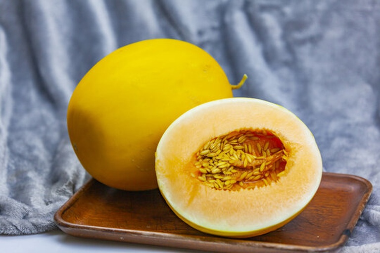 一张俯拍图,左侧是一个完整的金黄色哈密瓜,右侧是切开露出橙色果肉的半颗瓜,中间放置一个小型冰箱模型和一个显示24小时的时钟,传达‘保存时效’的主题,背景为浅色木纹桌面 完整的哈密瓜与切开的哈密瓜并列摆放,一侧有冰箱图标和时钟符号,象征保存与时间