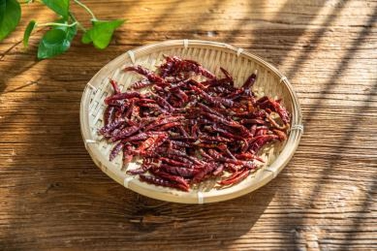 木质餐桌上陈列多种手工保存的尖椒制品:一串红色风干尖椒悬挂于左侧,中间是一玻璃罐浸泡在淡黄色液体中的腌制青椒,右侧为研磨好的红辣椒粉,背景为乡村厨房环境,自然光照射 风干与腌制的尖椒制品展示