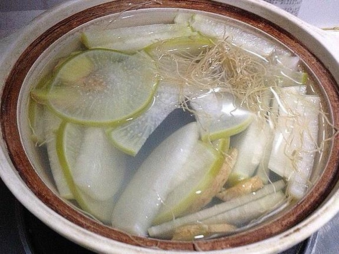 一位中年女性正在乡村厨房使用陶罐煎煮草本植物,锅中冒着蒸汽,旁边摆放着晒干的丝瓜根茎材料,台面上有剪刀、竹筛和玻璃瓶,整体氛围温馨自然 传统草药煎煮过程展示
