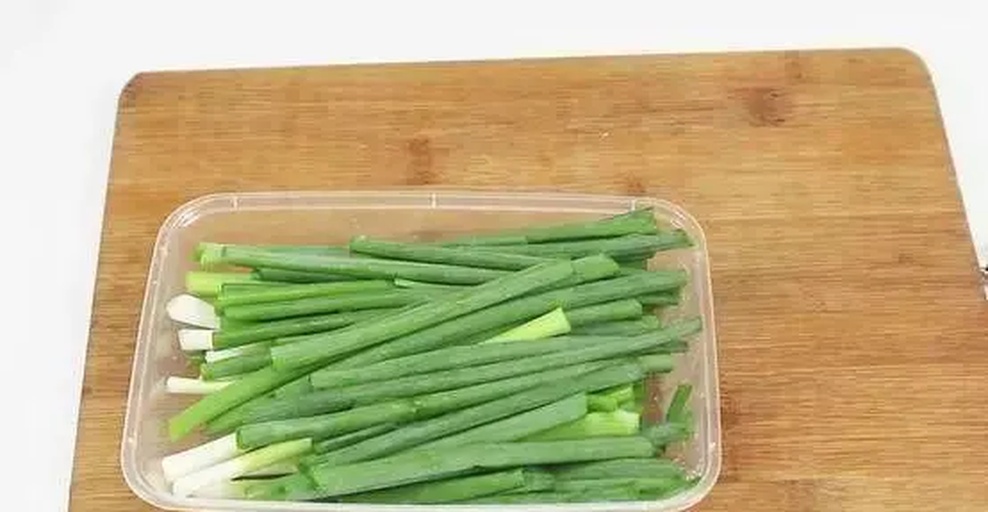 一个现代家庭厨房操作台上,左侧为用牛皮纸包裹竖立放置的大葱,中间为插入玻璃瓶中根部沾水的大葱,右侧为装在透明塑料盒内的切段葱花。背景可见调料架和燃气灶,体现真实生活场景 家庭厨房中大葱储存实景图