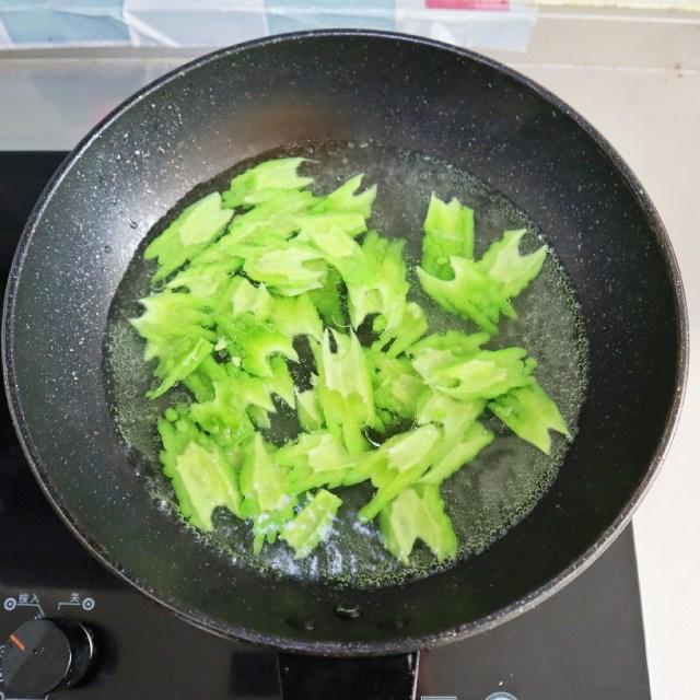 新鲜苦瓜斜刀切成薄片,正在倒入冒泡的沸水锅中进行焯水处理,锅边放有加盐的调料碗,背景为厨房灶台环境 苦瓜切片后放入沸水中焯烫的过程