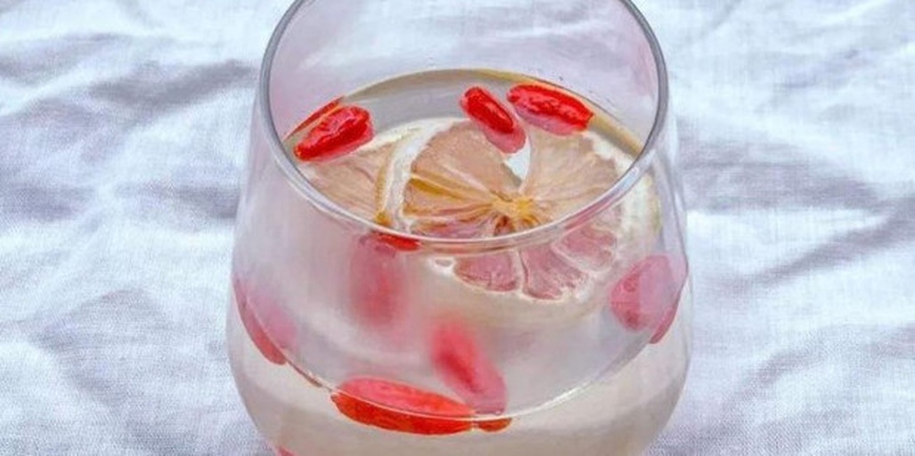 特写镜头展示一个装有混合冲泡饮品的透明水杯,顶部漂浮两片新鲜柠檬切片,中部悬浮几颗红润枸杞,底部可见细微絮状沉淀物。背景为切开的柠檬和干枸杞,强调食材组合带来的潜在不良反应 一个透明玻璃杯中装有混合液体,上层为橙黄色柠檬片漂浮,下层为红色枸杞沉底,中间有轻微分层和絮状物,象征化学反应发生