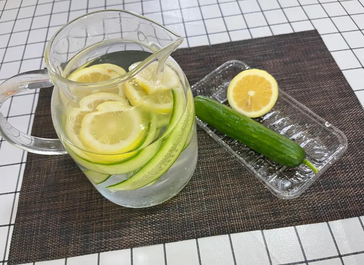 一杯用新鲜柠檬片泡制的淡黄色柠檬水,置于白色瓷盘上,旁边有半个切开的柠檬和一把小勺,展示家庭自制柠檬水的典型画面 柠檬切片浸泡在透明玻璃杯中的水里,背景为厨房台面,光线柔和,体现日常饮用场景