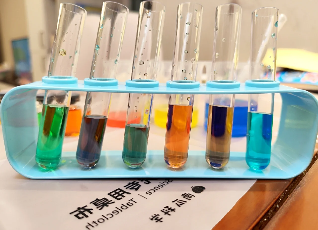 一组五个透明试管整齐排列在试管架上,分别装有紫甘蓝汁与不同物质混合后的液体,颜色从左至右依次为红色(酸性)、粉红、紫色(中性)、蓝色、黄绿色(碱性),背景为实验室风格桌面,标签清晰标注每种溶液类型 紫甘蓝汁在不同酸碱溶液中的颜色对比实验
