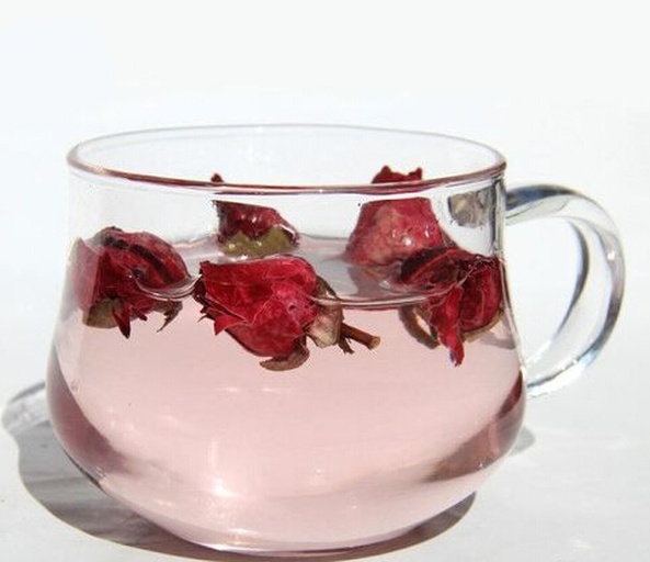 透明玻璃杯中正在冲泡玫瑰花茶,热水注入后花瓣缓缓展开,水中呈现淡粉红色,蒸汽袅袅上升,背景虚化的书本与笔记本电脑暗示办公场景 玫瑰花茶冲泡过程特写