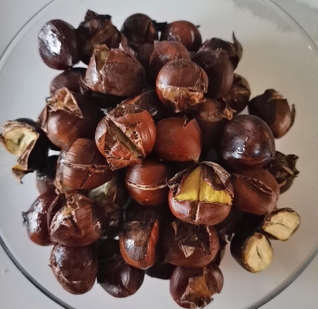 刚出炉的烤栗子特写：金棕色带光泽的带壳栗子堆叠在粗陶碗中，表面微泛油光，散发淡淡焦糖蒸汽，背景虚化为木质摊位和秋日暖阳