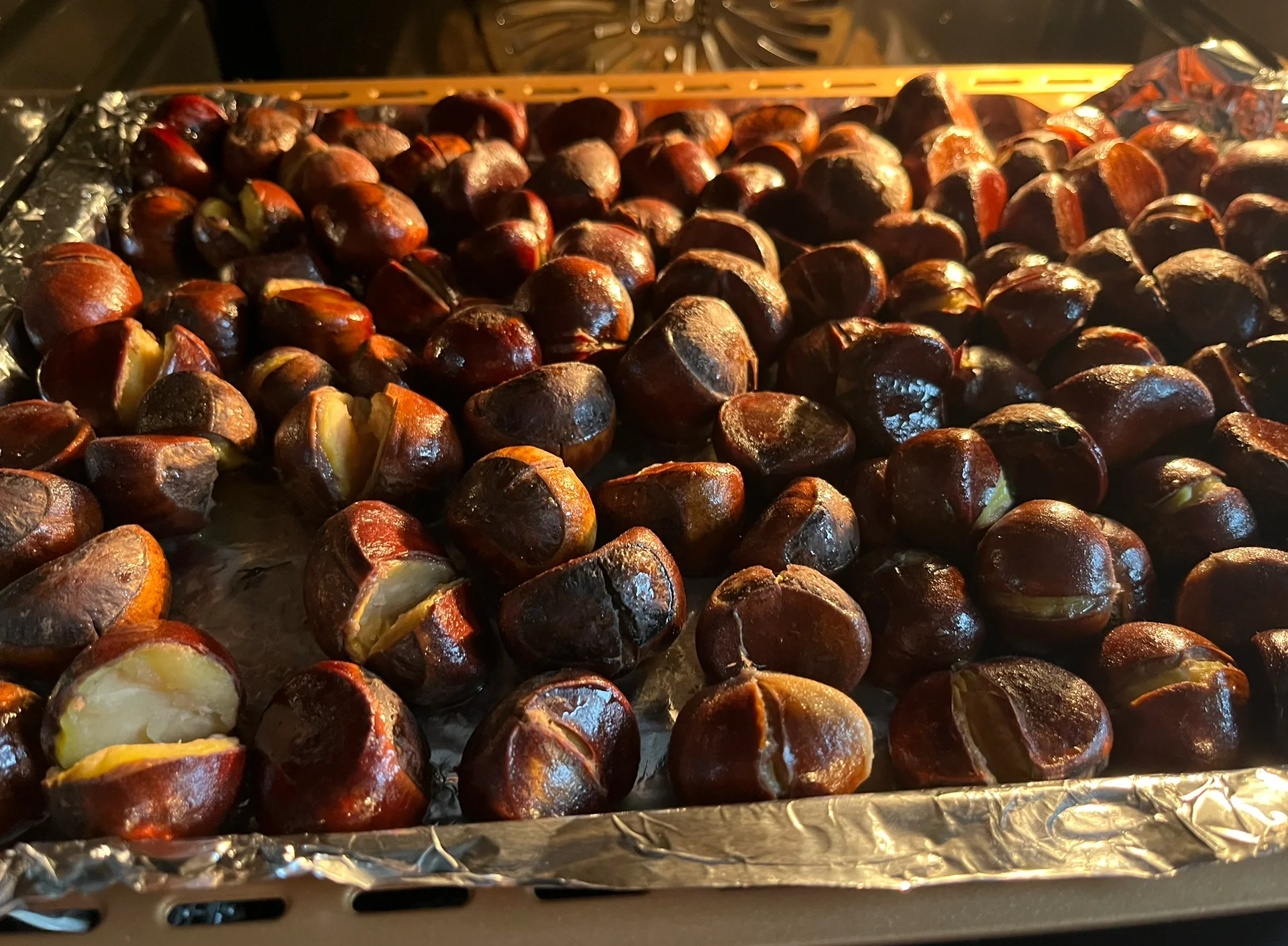 不锈钢烤盘上整齐排列着金黄色的油栗仁,正在烤箱中微微冒热气,背景可见温度计显示150摄氏度,旁边放置玻璃碗盛放剩余糖液,整体环境整洁明亮 油栗仁烘烤过程