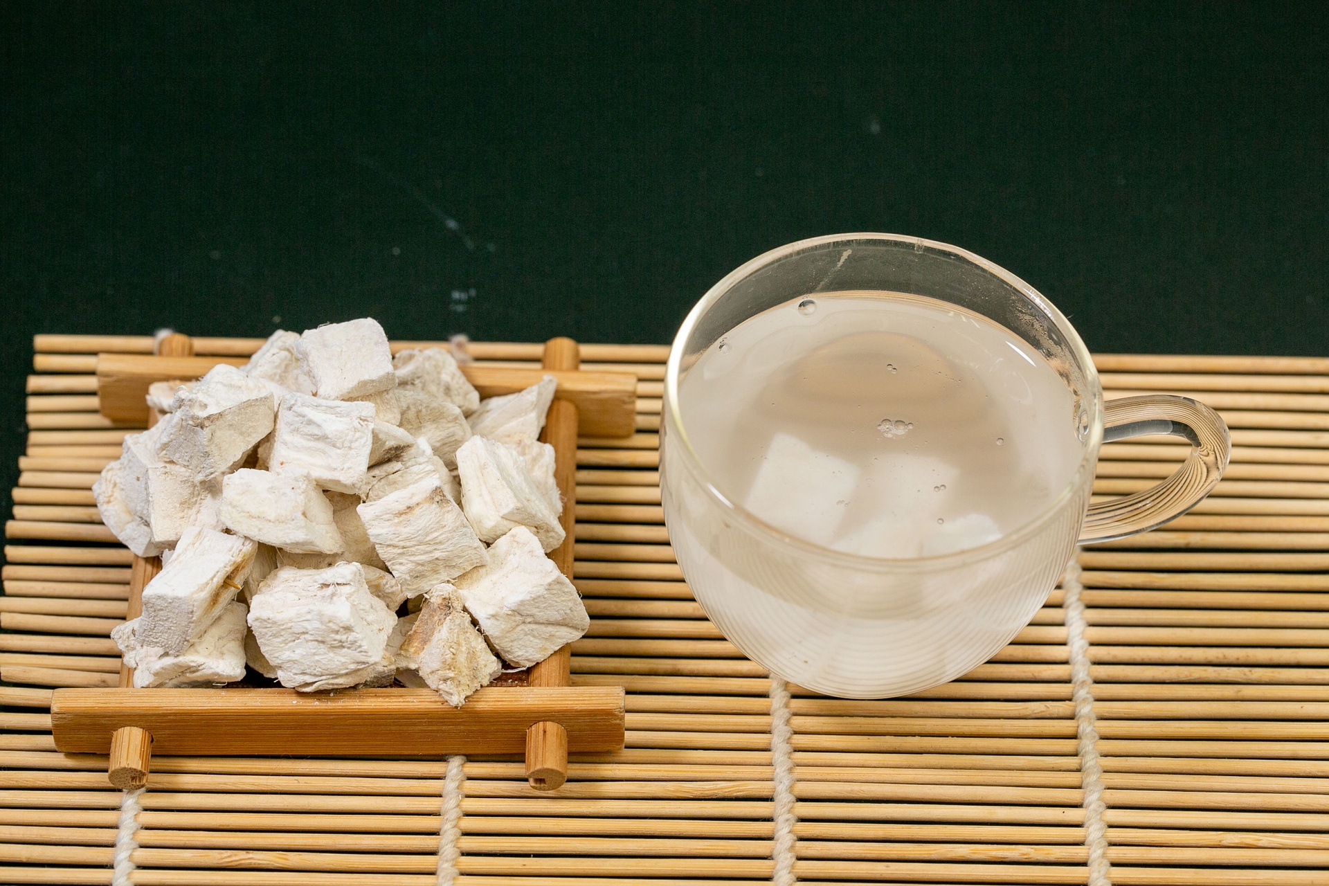 玻璃茶杯中正在冲泡白术片,热水倒入后呈现淡黄色茶汤,杯中有3片漂浮的白术片,旁边摆放着干燥的白术原片和陶瓷茶匙,桌面为浅色木纹背景 白术茶饮冲泡过程