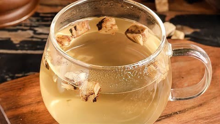 一张高清晰度的照片展示一杯透明玻璃杯中的粉葛茶,茶汤呈清澈的淡黄色,杯底可见几片浸泡后的粉葛薄片,杯子放置在木质茶盘上,背景虚化有茶具和绿植,营造出宁静雅致的饮茶氛围 粉葛茶冲泡成品展示