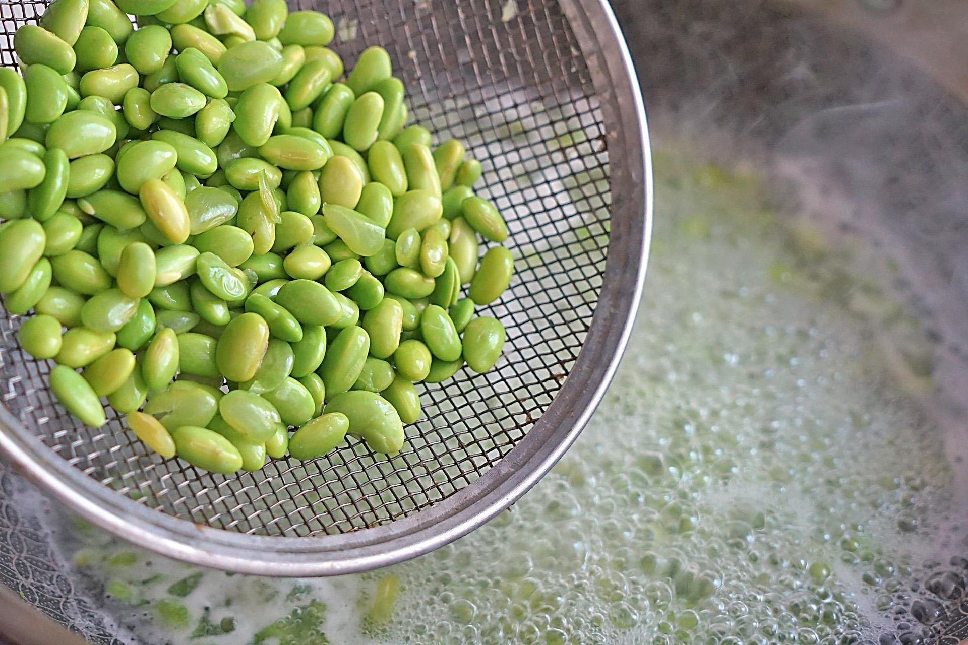 毛豆米焯水过程实拍,展示正确焯烫状态与冷却准备 锅中沸水翻滚,毛豆米正在焯烫,水面漂浮着绿色豆粒,旁边放有漏勺和冰水碗