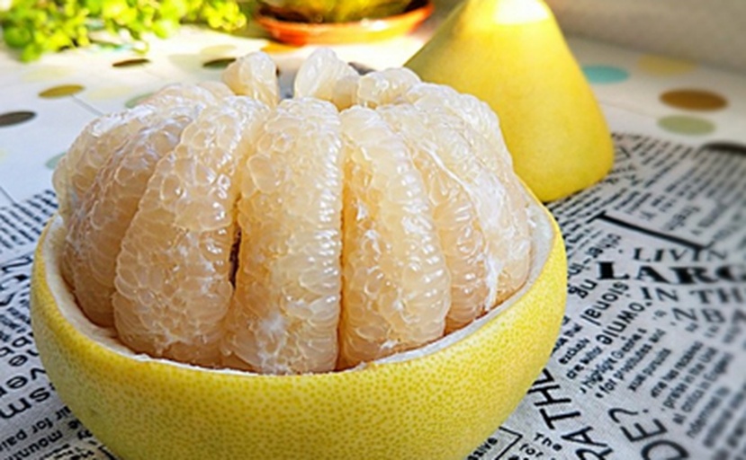 一个被横向切开的成熟柚子,露出晶莹剔透的粉白色果肉,汁水丰富,瓣膜清晰,周围点缀着几片绿叶,背景为木质餐桌,展现自然健康的食用场景 成熟的马来西亚本地柚子切开特写