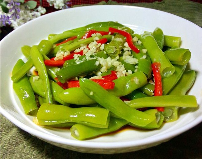 一盘刚出锅的蒜蓉清炒无筋豆盛放在蓝色瓷盘中，豆段斜切整齐，泛着油光，旁边点缀红椒丝