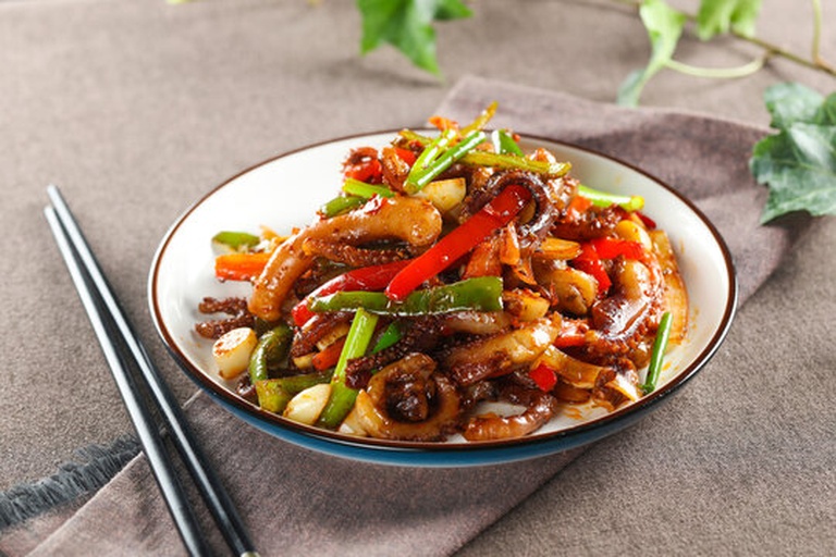 爆炒过程特写：铁锅中鱿鱼卷曲如麦穗，青红椒片鲜亮饱满，酱汁浓稠裹附食材，锅沿腾起白色蒸汽，火焰包裹锅底，呈现典型中式猛火爆炒动态场景
