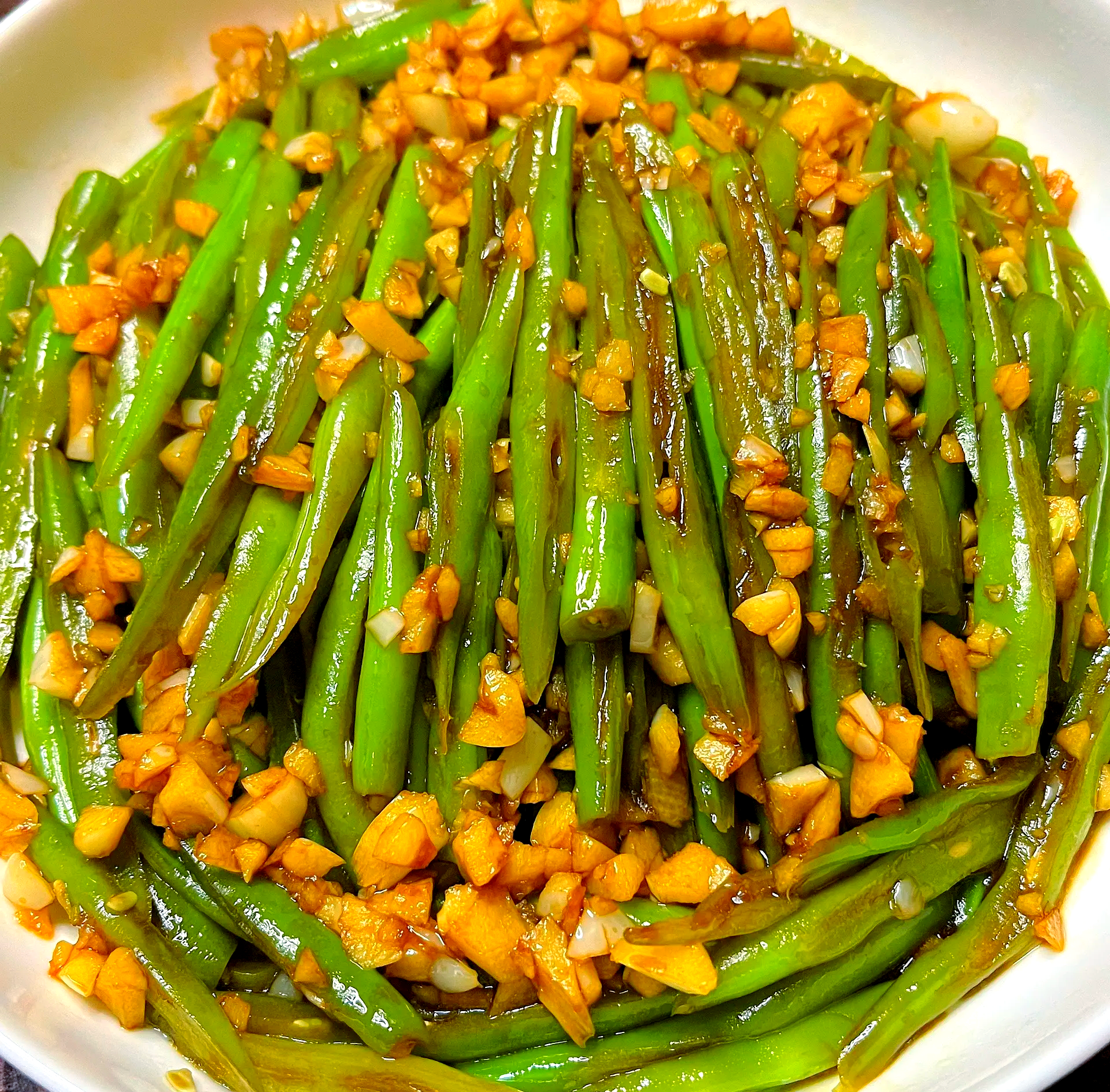 一张高清晰的餐桌摄影,盛放在白色瓷盘中的蒜蓉炒四季豆,色泽油亮翠绿,点缀金黄蒜粒,背景虚化,营造温馨家庭用餐氛围,突显菜肴美味与安全性 餐桌上一盘色香味俱全的蒜蓉炒四季豆成品图