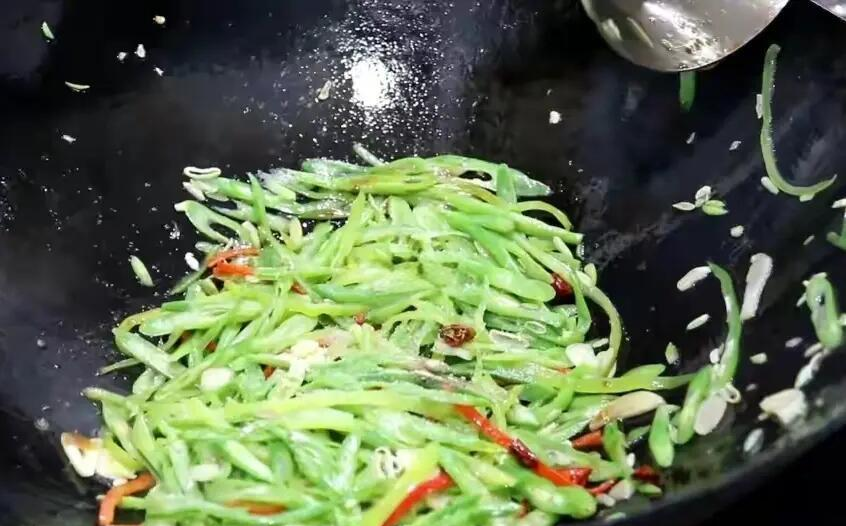 一个正在灶台上翻炒四季豆的家庭厨房场景,锅中为已变深绿色的四季豆段,搭配少量蒜片和红椒,厨师手持锅铲进行翻炒,炉火旺盛,显示高温烹饪状态 家庭厨房中炒制四季豆的过程