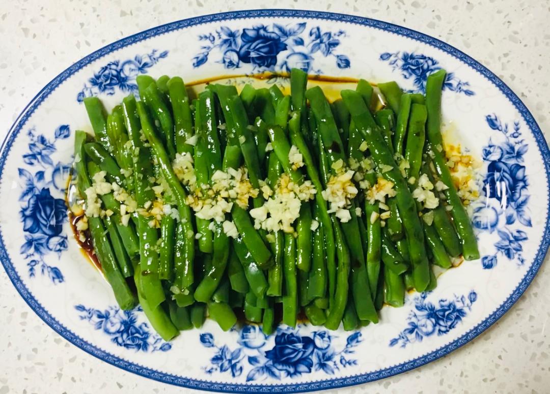 一盘刚出锅的蒜香焖豆角盛放在白色瓷盘中,豆角颜色油亮深绿,表面裹满浓郁酱汁,点缀着金黄蒜粒和红色辣椒圈,背景虚化的餐桌上配有米饭和筷子,展现诱人的家常美食氛围 成品豆角菜肴特写图