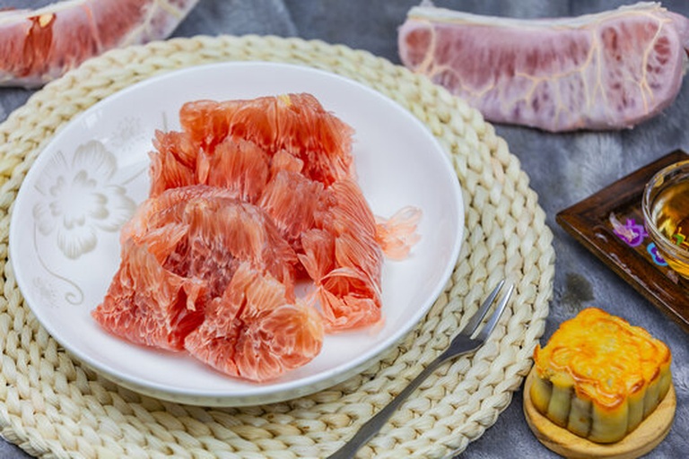 一张木质餐桌上摆放着一个被剥开的柚子,晶莹剔透的果粒清晰可见,旁边有一杯柚子茶和一小碟果肉,营造出温馨的家庭享用场景 家庭中剥开的柚子特写