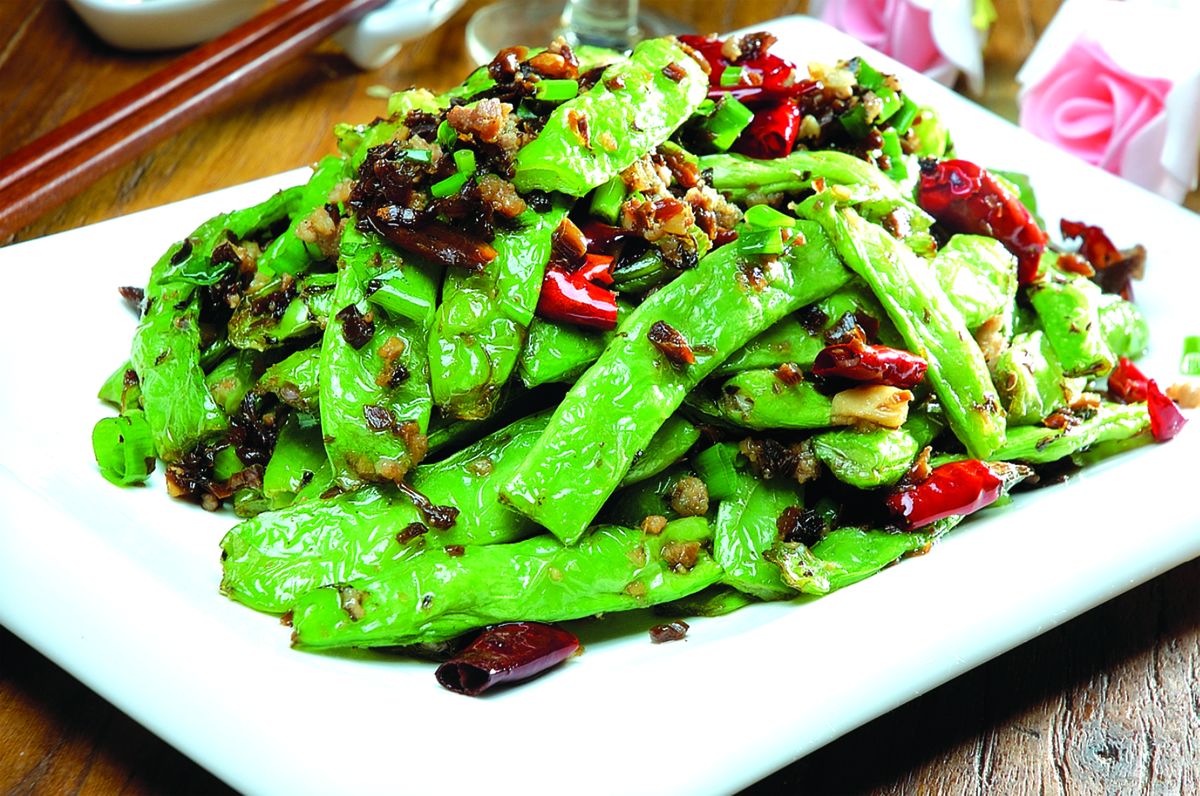 成品四季豆特写：一盘油亮翠绿带微黄的四季豆盛于白瓷盘中，豆角段整齐微卷，表面可见细密虎皮纹，撒有金黄蒜末与红椒丝，背景虚化显示中式灶台与铁锅