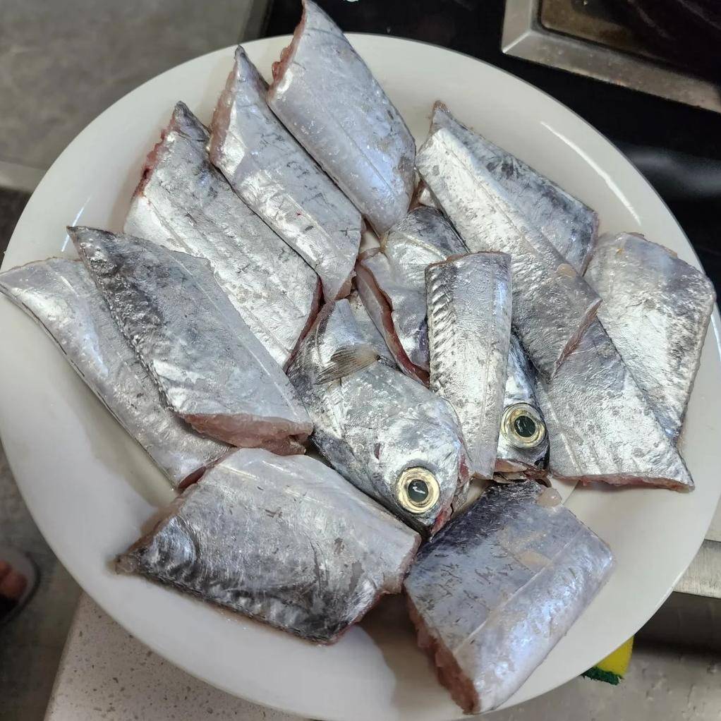 一名厨师正在操作台上用刀清理一条新鲜带鱼,去除内脏和银色表皮,旁边摆放着切好的姜片和葱段,案板整洁,光线明亮 带鱼清洗与切段过程