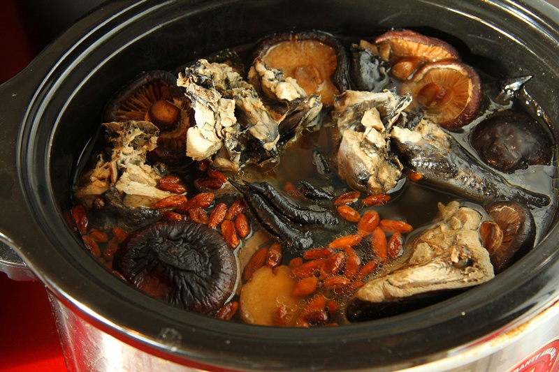 高清俯拍:砂锅中炖煮中的乌鸡煲花旗参药膳,可见完整乌鸡块、切片花旗参(米黄色半透明薄片)、枸杞子与少量陈皮丝,汤色清亮微黄,表面浮有细小油珠,无浑浊沉淀 乌鸡煲花旗参药膳特写