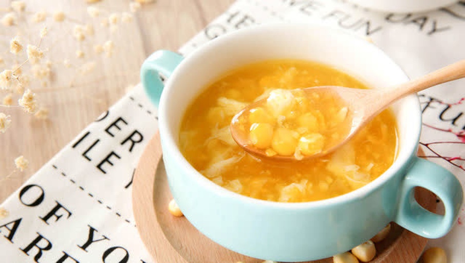 奶白色糯玉米粥盛在瓷碗中，冒着热气，旁边摆放木勺和玉米棒