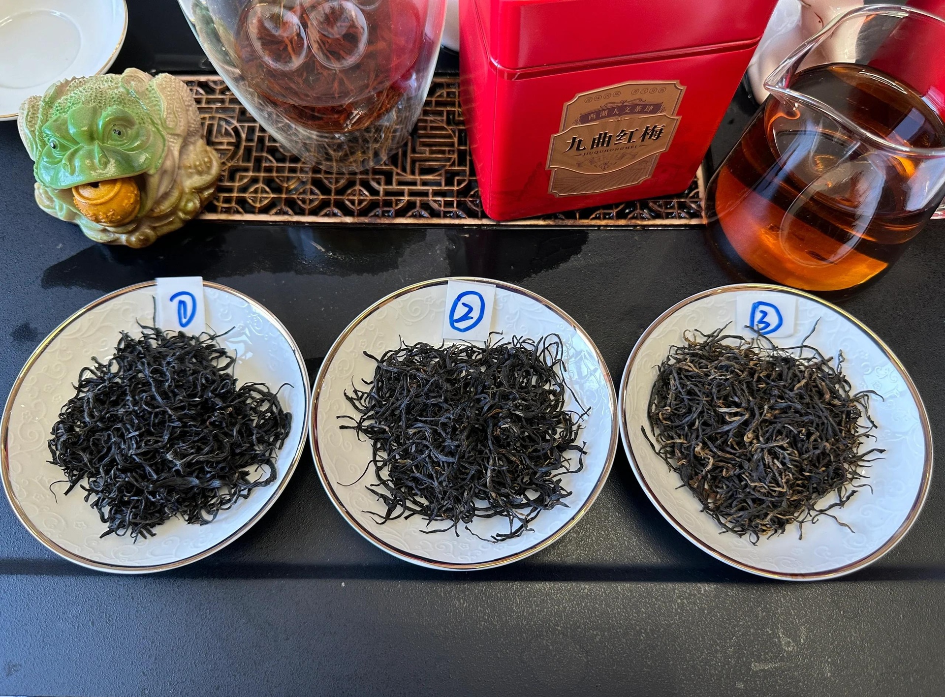 干茶呈深褐色,条索紧细卷曲如鱼钩,表面油润有光泽,均匀整齐地铺展在白色瓷盘上,背景为淡木色桌面,展现传统工夫红茶的精致外形 九曲红梅干茶特写