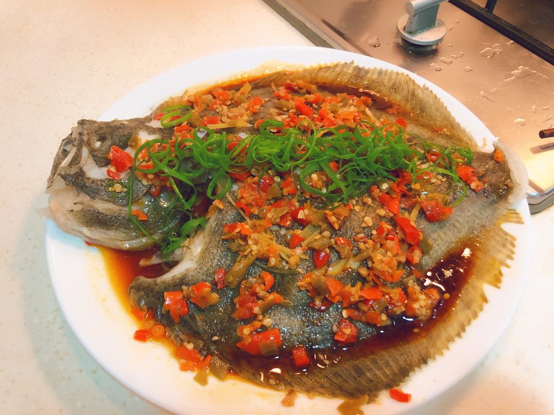 一整条清蒸大菱鲆摆放在白色瓷盘中,鱼身完整未断裂,表面淋有透明豉油汁,点缀姜丝与葱段,热气升腾,鱼肉呈蒜瓣状自然裂开,显示出良好的熟成状态,皮面光亮富有弹性 清蒸多宝鱼成品特写