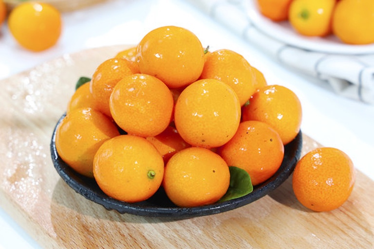 一盘新鲜采摘的金黄色金桔,散落在木质桌面上,阳光照射下果皮晶莹剔透,部分果实带有绿叶,背景为模糊的果园环境 金桔在阳光下的特写镜头