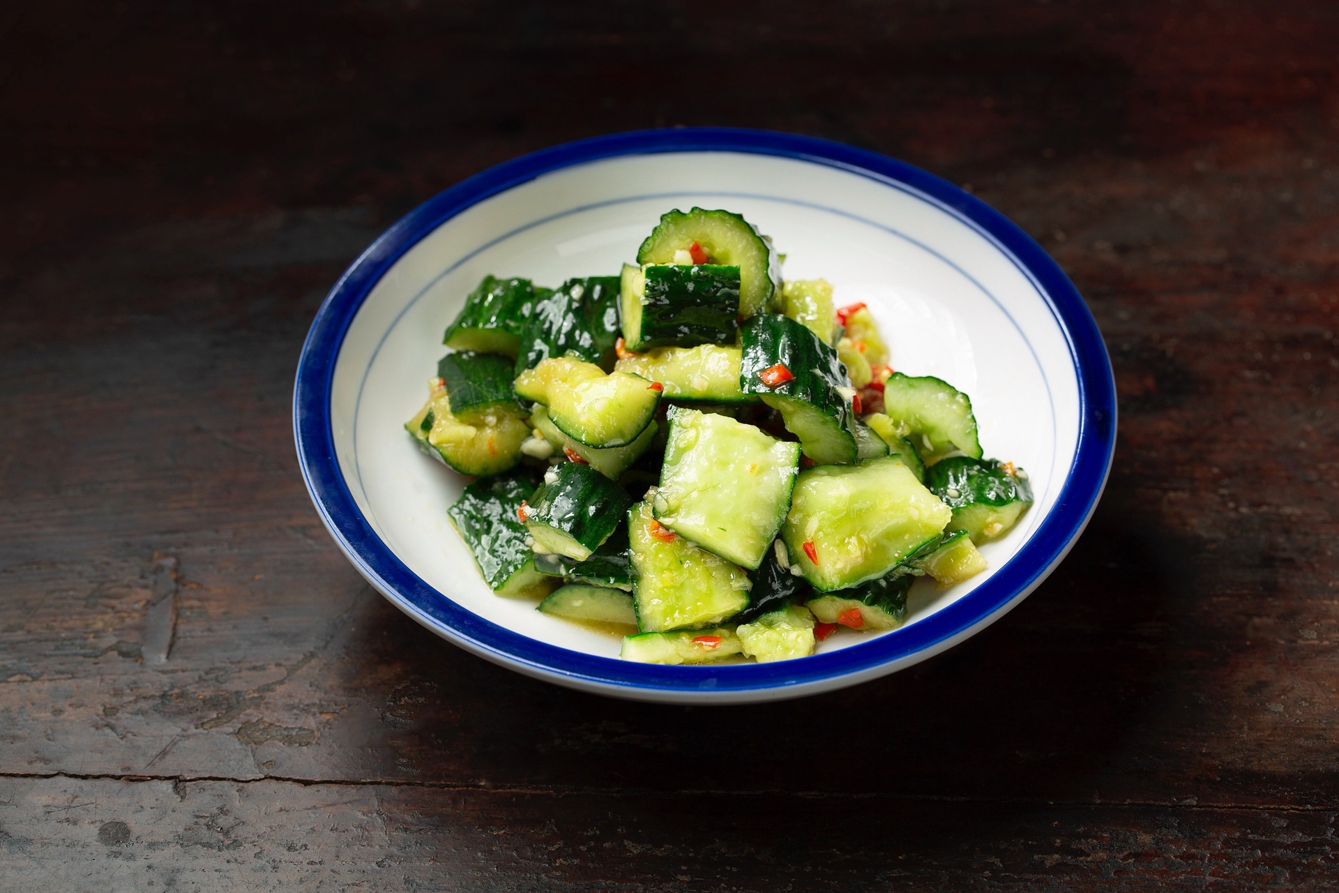 一盘晶莹剔透的凉拌小黄瓜摆放在白色瓷盘中,黄瓜呈浅绿色裂纹状,表面泛着香油光泽,点缀着细碎蒜末和红色小米辣圈,背景虚化的竹筷正夹起一段,展现其脆嫩质地 凉拌小黄瓜成品特写