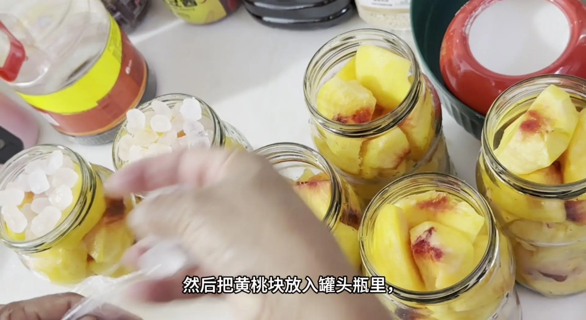 厨房台面上摆放着玻璃罐装的新鲜黄桃块、一瓶打开的原味酸奶、一个搅拌碗和一把不锈钢勺,正在将黄桃倒入酸奶中进行混合,展示家庭自制黄桃酸奶的过程,强调食材透明与手工制作的安全感 黄桃酸奶制作过程示意图