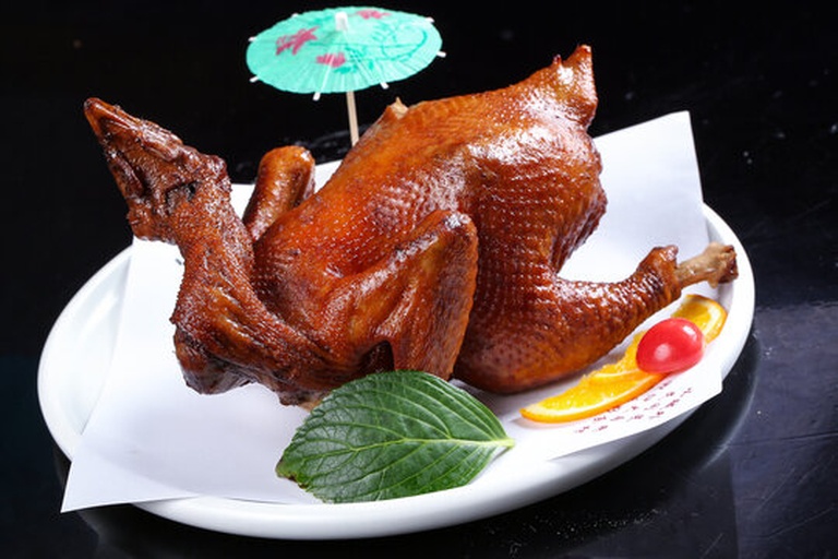 一只色泽金黄油亮的烤鸡放置在白色瓷盘中,鸡皮酥脆反光,周围摆放着焦糖化的橙片,背景虚化可见烤箱和厨房环境,整体呈现温暖诱人的晚餐氛围 橙香烤鸡成品特写