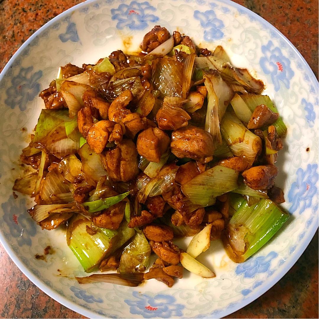 一盘刚出锅的葱爆鸡腿盛放在白色圆盘中,鸡丁红润油亮,大葱段分布均匀,表面泛着光泽,背景虚化的餐桌上配有米饭和筷子,营造温馨家常氛围 成品葱爆鸡腿特写