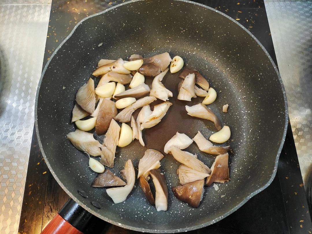 厨房操作台上的炒锅正在翻炒平菇，锅中平菇呈金黄色，表面裹着晶莹酱汁，旁边摆放着切好的蒜末、葱段和调味碗