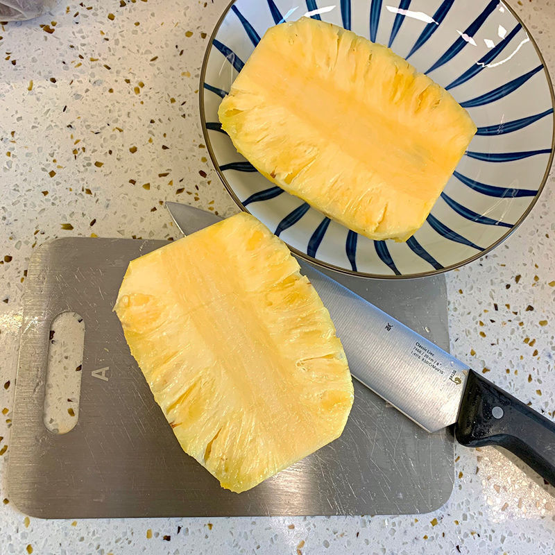 一个完整的菠萝被横向切开,展示其内部金黄色的致密果肉,切面湿润有光泽,周围摆放着几块去皮切块的菠萝,背景为木质砧板和厨房环境,体现家庭食用场景 切开的菠萝特写,露出金黄色的果肉,汁水丰富,旁边有一把水果刀和几块已切好的菠萝块