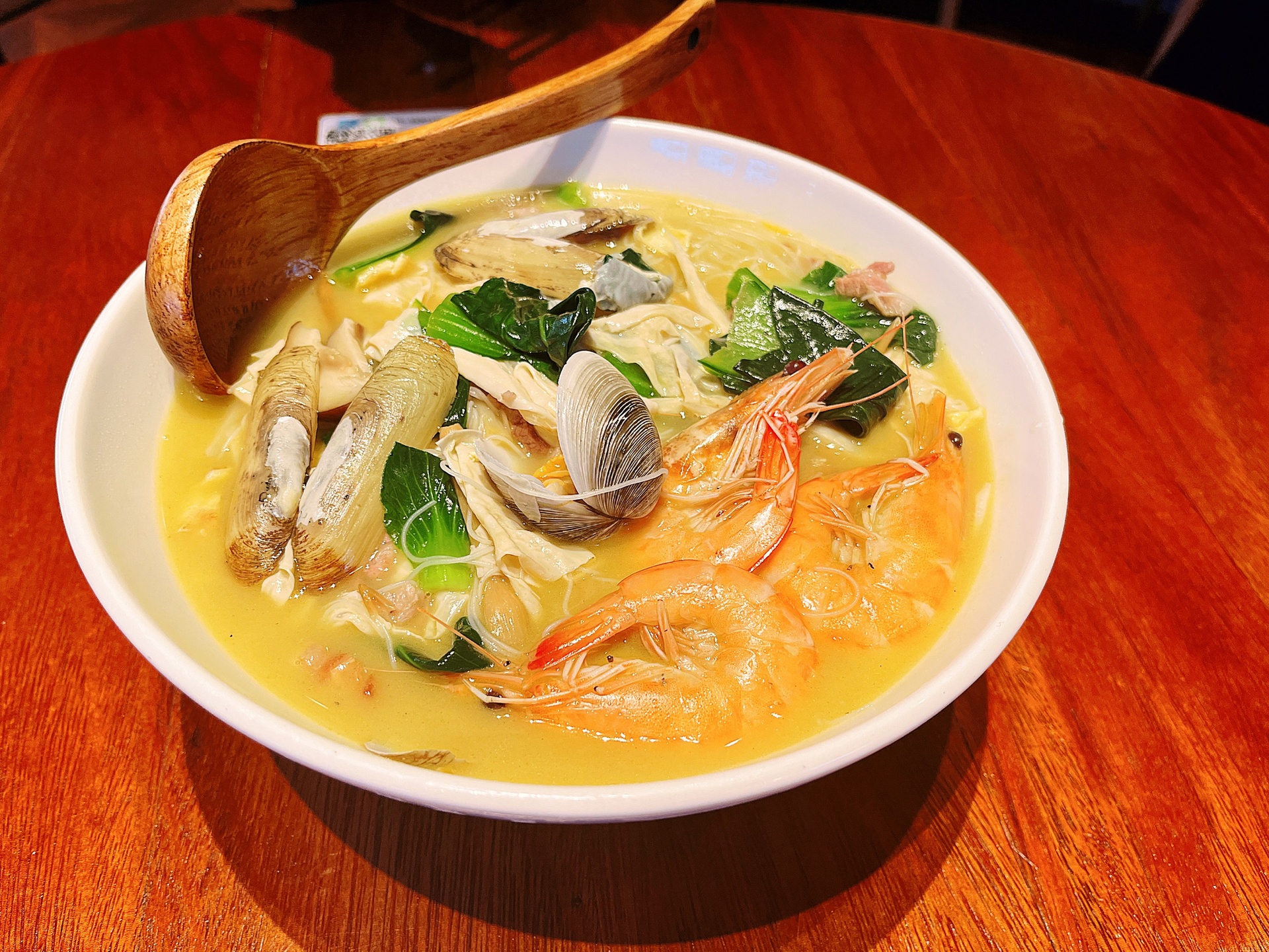 一碗冒着热气的手工拉面,汤色微黄清澈,表面浮着几片薄切的新鲜面姜,点缀葱花和少许香菜。面条整齐堆叠,旁边放有一双木筷和一个小瓷碟,背景为原木餐桌,环境温馨居家 一碗热气腾腾的姜汤面