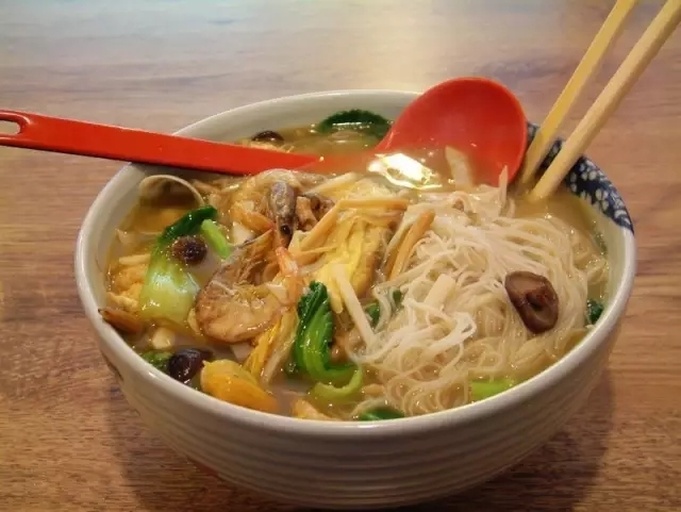 一碗盛满的海鲜姜汤面,汤色金黄清澈,面条整齐铺底,上面覆盖着红润的虾仁、张口的蛤蜊、乳白的鱼片和浅褐的鱿鱼圈,姜片隐约可见,撒有翠绿香菜和葱花,背景虚化体现食欲感 成品海鲜姜汤面特写