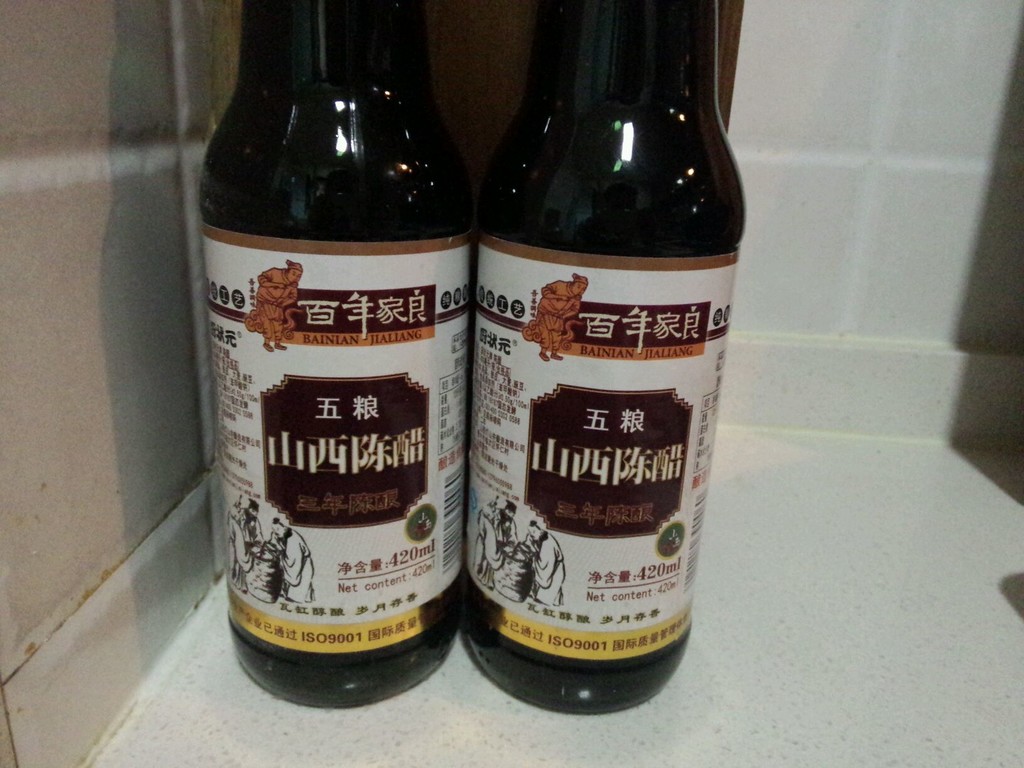三款合规酿造醋并排摆放：左为山西老陈醋陶罐装，中为镇江香醋玻璃瓶，右为纯粮米醋方形瓶，瓶身均带SC标志与执行标准号，背景为木质砧板