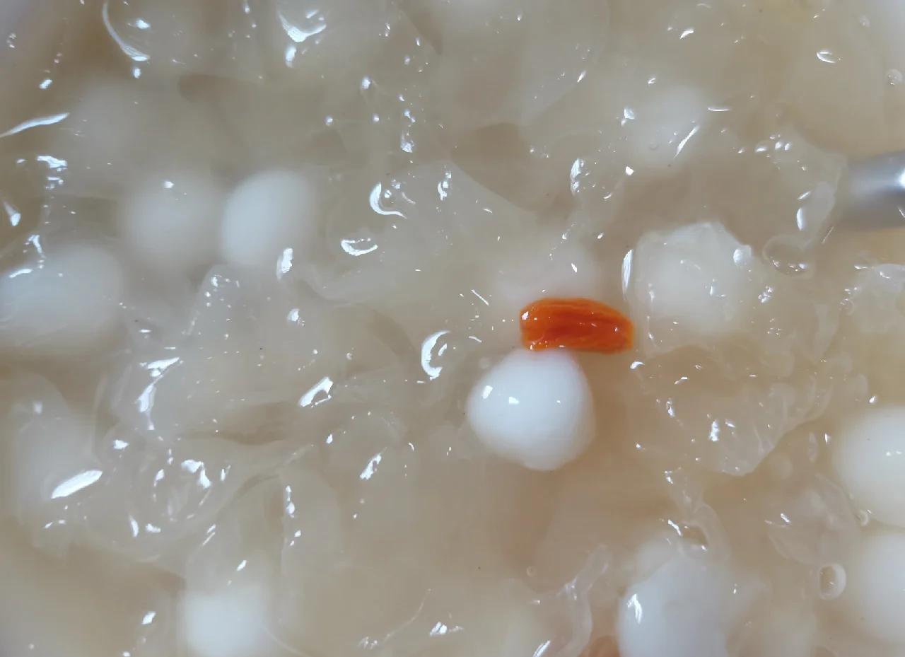 一碗盛满的菠萝银耳甜汤,透明银耳胶质丰富,菠萝块呈嫩黄色,枸杞点缀其间,汤体清澈微稠,置于木质餐桌,背景有蒸汽袅袅升起 菠萝银耳甜汤特写