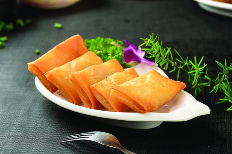 一张木质餐桌上摆放着三个小碟子:左侧是卷好的米纸春卷切段,露出内部橙色脆柿条和蔬菜;中间是一杯粉色冰沙插着吸管;右侧是方形披萨一角,上面铺有黄橙色脆柿片、绿色芝麻菜和白色奶酪碎,整体色彩鲜艳诱人 脆柿创意料理拼盘