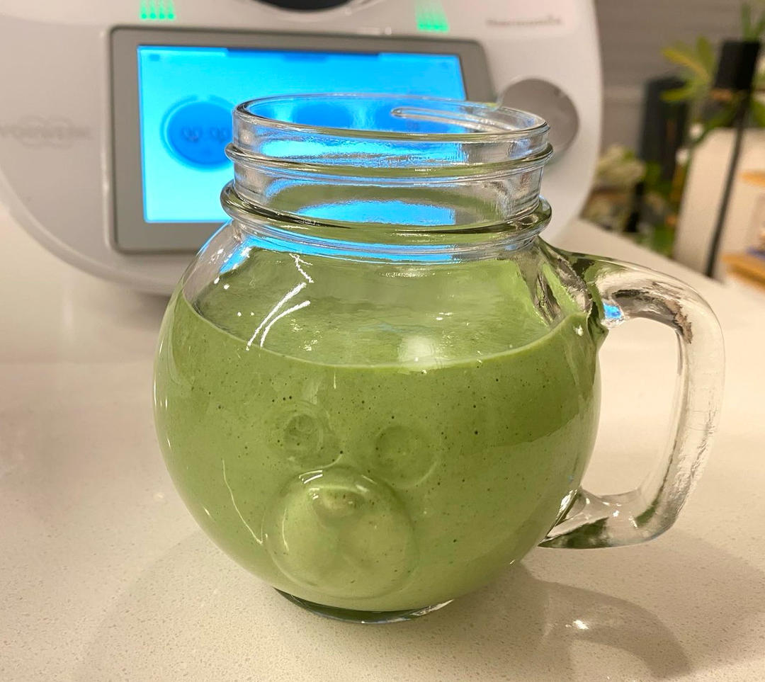 一杯盛满绿色奶昔的透明玻璃杯，杯壁凝结水珠，旁边放一把金属长勺，背景虚化的厨房中有搅拌机和食材残余