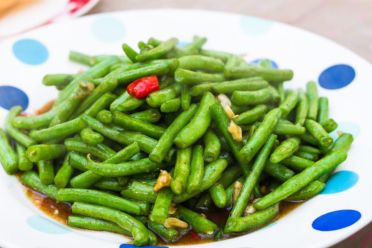 一盘刚出锅的短豆角炒土豆盛放在白色瓷盘中,土豆呈金黄色,短豆角翠绿鲜亮,点缀着金黄蒜片和红色干辣椒段,整体油润有光泽,背景为木质餐桌,营造温馨家常氛围 成品短豆角炒土豆装盘效果