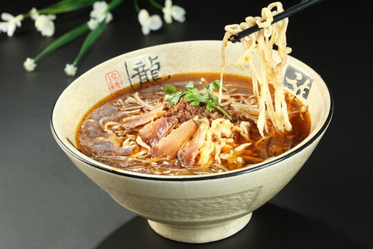 一碗手工面条上淋满红亮辣椒油，撒有葱花和芝麻，旁边摆放木筷和辣椒罐