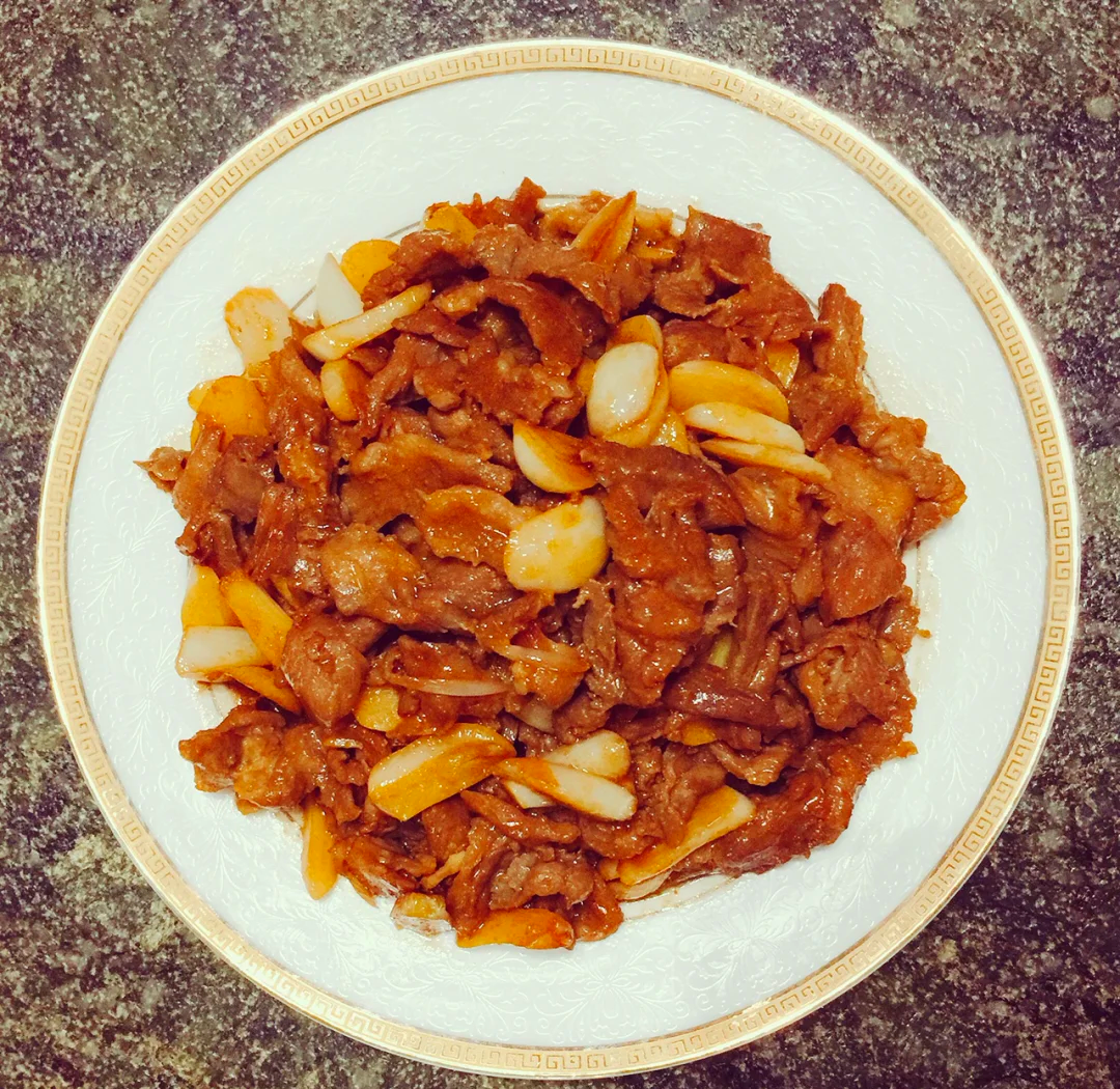 成品俯拍：青红椒丝围边，中间堆叠油亮诱人的蒜香牛肉片，表面隐约可见金黄蒜粒与翠绿葱花，盘底有少量琥珀色酱汁反光