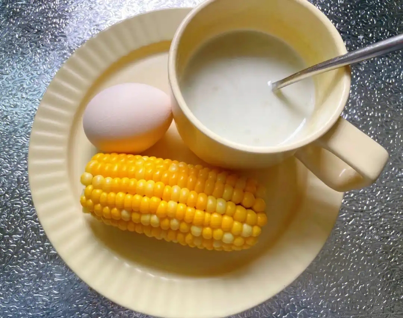 一位母亲在明亮整洁的现代厨房中用玉米油煎蛋,锅具上方有轻微油烟但不明显,桌面上摆放着一瓶透明包装的玉米油,标签清晰可见,旁边配有新鲜玉米棒和蔬菜,展现健康饮食的生活方式 家庭厨房中使用玉米油烹饪的场景