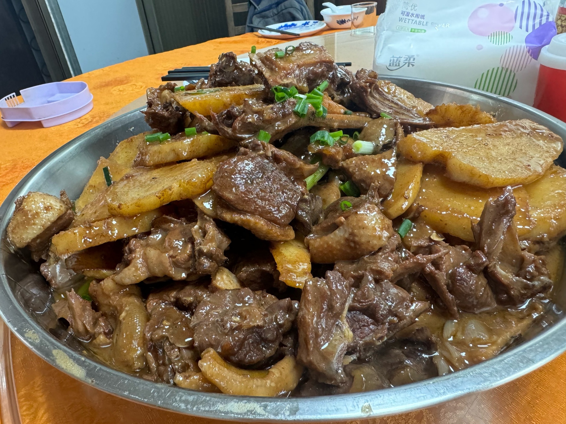 一道装在白色瓷盘中的红烧鸡翅炖土豆,鸡翅色泽红亮,土豆金黄软糯,表面覆盖着浓稠油亮的酱汁,旁边配有几根翠绿葱花点缀,背景为简约餐桌布景 成品红烧鸡翅炖土豆装盘效果图