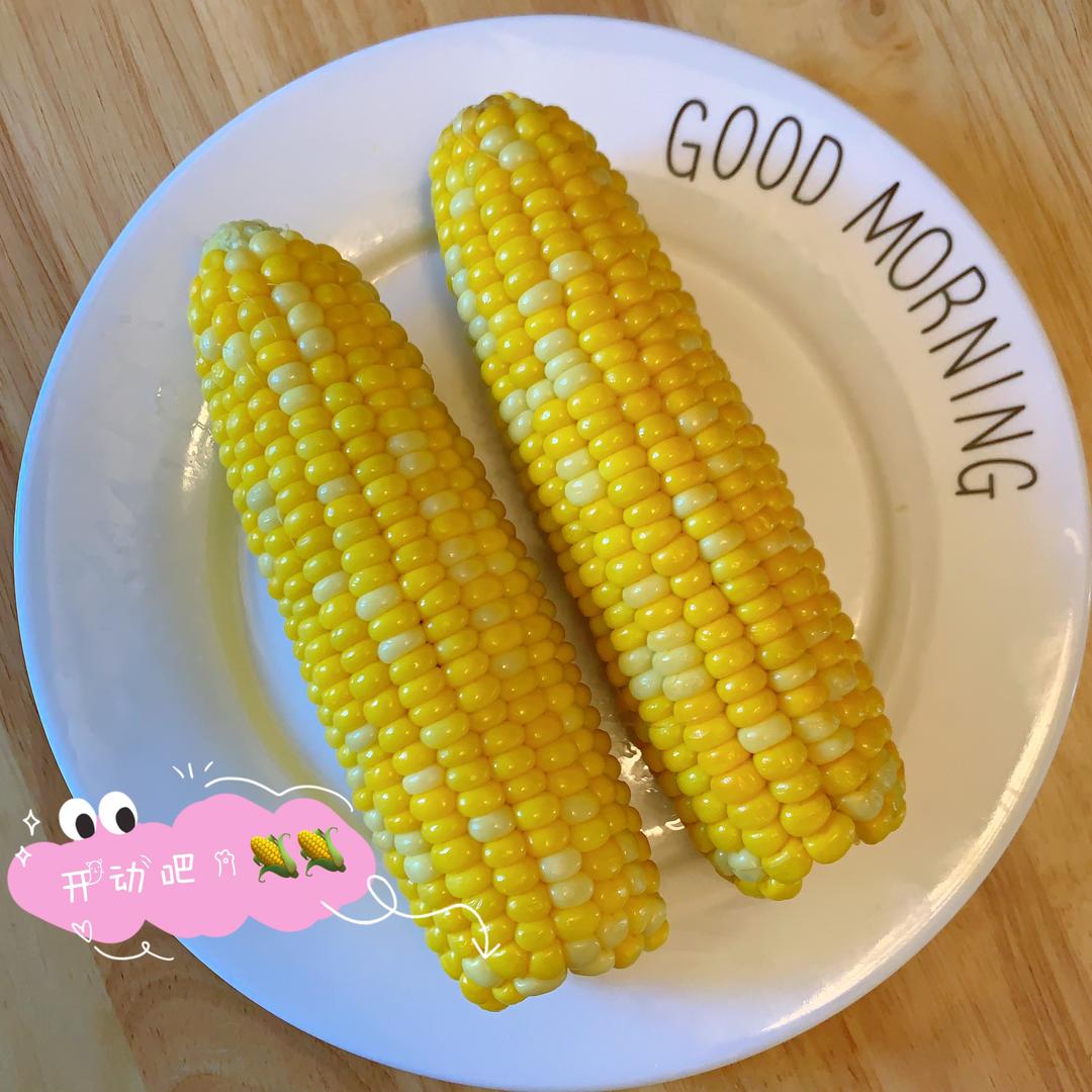 一家三口在餐桌前共享早餐,母亲正在将一根蒸熟的糯玉米切成小段放入儿童餐盘中,孩子笑脸迎接,桌上还有牛奶杯和水果,营造温馨健康的饮食氛围 家庭亲子共食糯玉米场景