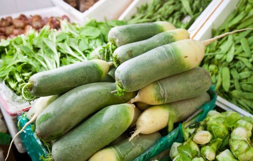 一个露天菜市场的摊位,摆放着大量新鲜的青萝卜,整齐排列在竹筐中,旁边有其他蔬菜如白菜、胡萝卜,商贩正在整理货物,背景为清晨阳光下的街道 菜市场摊位上的青萝卜