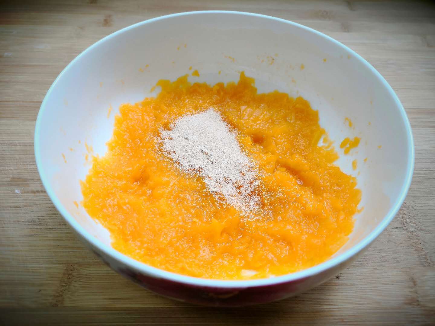 一个白色陶瓷碗中装有橙黄色的南瓜泥,旁边撒着一些糯米粉,一只手正在将两者混合揉合成光滑的黄色面团,桌面上还有剩余的糯米粉和一把小刮刀 南瓜泥与糯米粉混合揉团过程